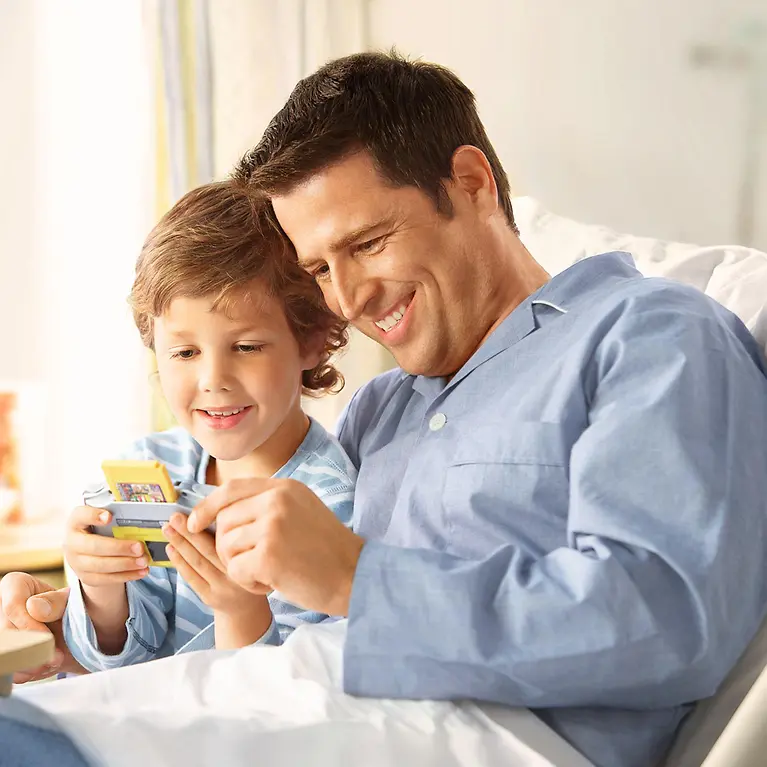 Ein Mann und ein Kind sitzen in einem Krankenbett und schauen zusammen auf einen Gameboy.
